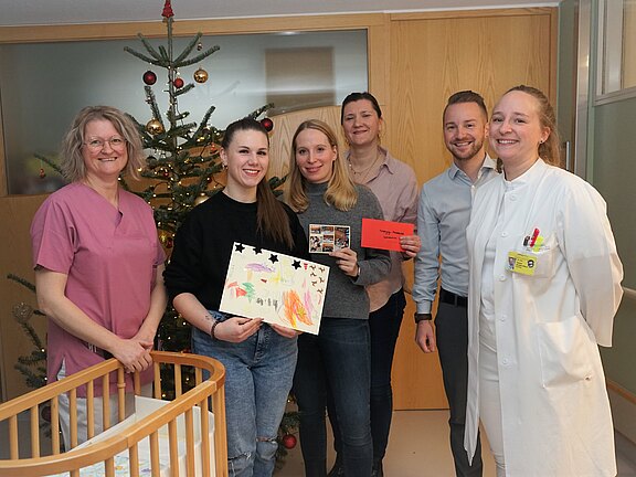 Gruppe lächelnder Menschen in einem Krankenhaus, neben einem Weihnachtsbaum. Zwei Personen halten eine Kinderzeichnung und eine rote Karte oder Umschlag. Pflege- und Ärzteteam posiert gemeinsam, bei einer Übergabe oder Spendenaktion auf der Kinderstation.