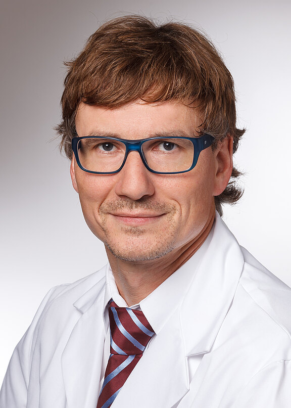 Porträt eines erwachsenen Mannes mit kurzer brauner Frisur und blauer Brille, in weißem Kittel und gestreifter Krawatte, vor neutralem Hintergrund, blickt freundlich in die Kamera.