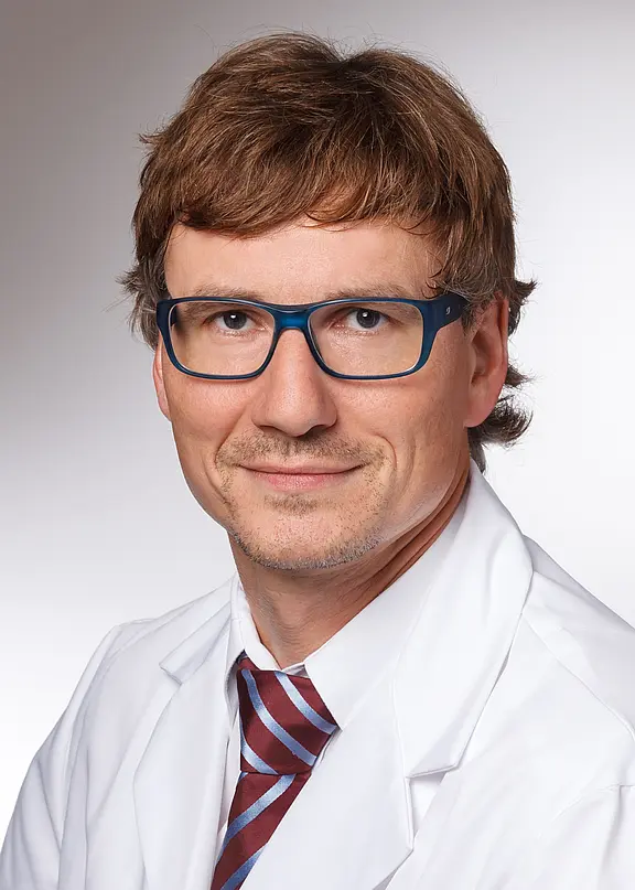 Porträt eines erwachsenen Mannes mit kurzer brauner Frisur und blauer Brille, in weißem Kittel und gestreifter Krawatte, vor neutralem Hintergrund, blickt freundlich in die Kamera.
