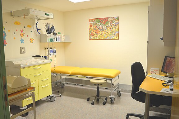 Heller Untersuchungsraum einer Kinderarztpraxis: gelbe Untersuchungsliege, Wickelkommode mit Schubladen, Wandregal mit medizinischen Geräten, Schreibtisch mit Computer. Bunte Wanddeko schafft eine kinderfreundliche Atmosphäre.