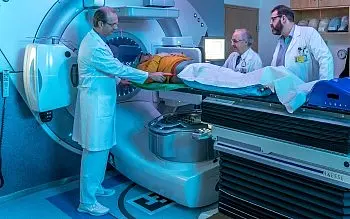 Drei Mediziner in weißen Kitteln positionieren eine liegende Person auf einer Trage vor einem großen runden medizinischen Gerät, ähnlich einem MRT oder Strahlentherapie-System. Sie überprüfen die Lage und bereiten die Untersuchung vor.