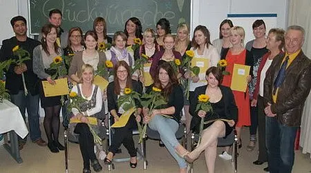 Eine Gruppe von 20 Personen, darunter Frauen und Männer, steht und sitzt zusammen in einem Raum. Alle halten Sonnenblumen und einige haben Glückwunschkarten in den Händen. Im Hintergrund ist eine Tafel mit der Aufschrift „Glückwünsche“ sichtbar.