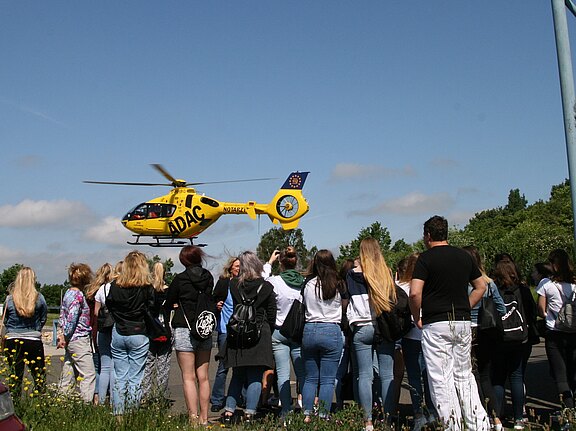 Ein gelber ADAC-Rettungshubschrauber mit Notarzt-Aufschrift schwebt knapp über dem Boden; eine große Gruppe Zuschauer beobachtet die Landung auf einer offenen Fläche bei klarem Himmel.