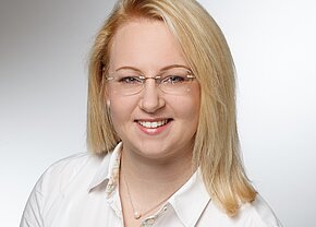 Barbara Bär Porträt einer lächelnden Person mit blonden Haaren und einer weißen Bluse. Sie trägt eine dezente Kette mit einer Perle und eine Brille. Der Hintergrund ist hell und neutral, wodurch der Fokus auf dem Gesicht liegt.