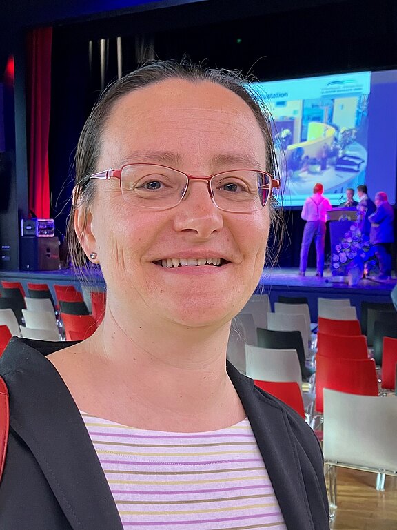 Porträt einer lächelnden Person mit Brille in einem Veranstaltungssaal. Hinter ihr stehen viele leere Stuhlreihen; auf der Bühne ist eine kleine Gruppe vor einer großen Präsentationsfolie zu sehen.