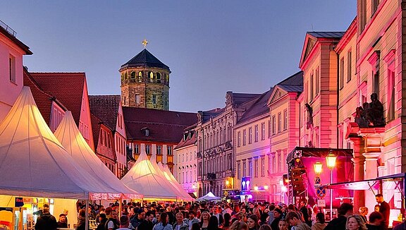 Belebte Stadtstraße bei Abendlicht, gesäumt von historischen Gebäuden und Festzelten. Viele Menschen genießen eine Veranstaltung. Im Hintergrund steht ein Turm mit einer goldenen Kreuzspitze. Die Lichter verleihen der Szene eine festliche Atmosphäre.