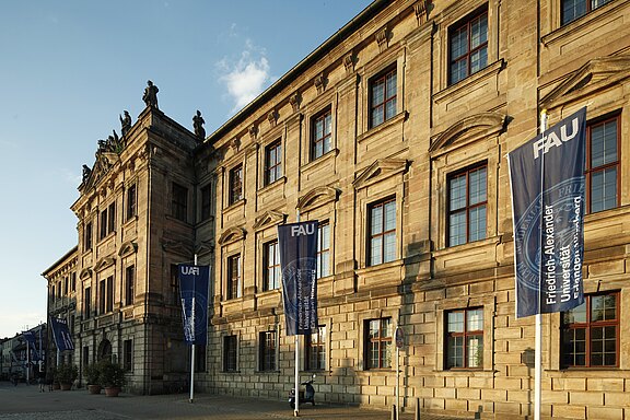 Historische Sandsteinfassade eines barocken Universitätsgebäudes im Abendlicht. Mehrere blaue Fahnen mit dem Schriftzug „FAU – Friedrich-Alexander-Universität Erlangen-Nürnberg“ säumen den Gehweg vor der Fassade. Große rechteckige Fenster und Dachfiguren prägen die Architektur.