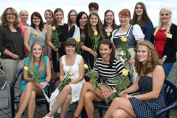 Eine Gruppe von 15 Frauen, die fröhlich lächeln, posieren für ein Foto. Einige halten gelbe Rosen in den Händen. Sie tragen verschiedene Kleider, sowohl festlich als auch casual, und stehen in einer Reihe sowie sitzen auf Stühlen. Der Hintergrund zeigt einen hellen Himmel.