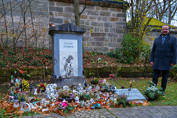 Ein Mann steht neben einem Gedenkort mit einer Stele, auf der "U ns e r e n K i n d e r n" geschrieben steht. Rund um die Stele sind zahlreiche Blumen, kleine Figuren und persönliche Andenken platziert, die dem Andenken an verstorbene Kinder gewidmet sind.