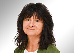 Siglinde Hübner Porträt einer lächelnden Frau mit schulterlangem, dunklem Haar. Sie trägt einen grünen Blazer über einem weißen Oberteil vor neutralem Hintergrund.