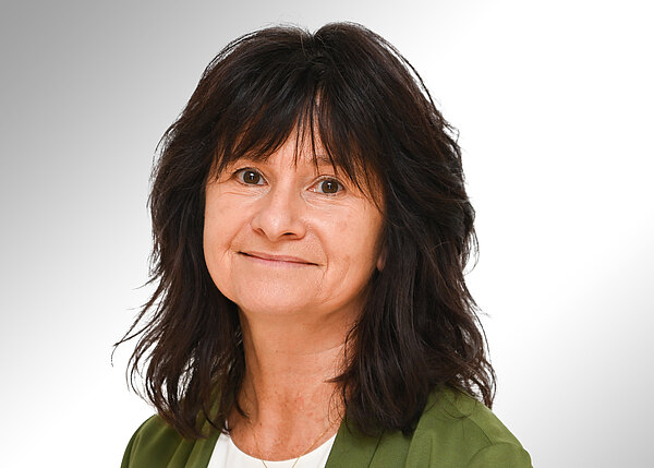 Porträt einer lächelnden Frau mit schulterlangem, dunklem Haar. Sie trägt einen grünen Blazer über einem weißen Oberteil vor neutralem Hintergrund.