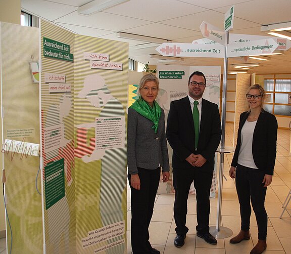 Drei Personen stehen vor einer Infowand in einem hellen Raum. Plakate und ein Wegweiser thematisieren Arbeit: ausreichende Zeit, verlässliche Bedingungen und Qualität. Die Gruppe präsentiert die Ausstellung und lächelt in die Kamera.