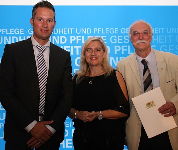 Drei Personen in formeller Kleidung posieren vor blauem Hintergrund mit der Aufschrift „Gesundheit und Pflege“. Der Mann rechts hält eine Urkunde mit Wappen, die Frau in der Mitte lächelt, der Mann links steht mit gefalteten Händen.