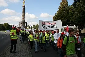 Eine Gruppe von Menschen in leuchtenden T-Shirts marschiert auf einer Straße und trägt ein großes Banner mit der Aufschrift "Klinikum Bayreuth". Im Hintergrund ist ein Monument zu sehen. Die Atmosphäre wirkt demonstrativ und engagiert.