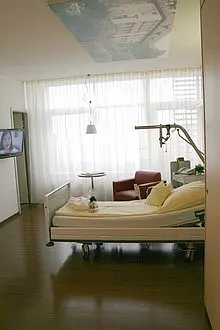 Ein Krankenhauszimmer mit einem elektrisch einstellbaren Bett, gelben Laken und einem Nachttisch. Neben dem Bett steht ein Tisch mit einer Wasserflasche. In der Ecke befindet sich ein roter Sessel und an der Wand ein Fernseher. Große Fenster sorgen für viel Licht.