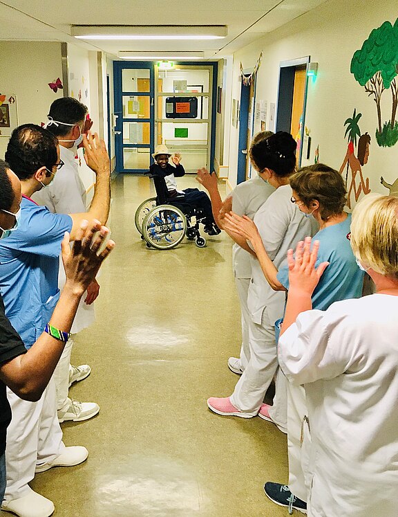 In einem Krankenhausflur bildet Pflegepersonal eine Gasse und klatscht einem Kind im Rollstuhl zu, das den Flur entlangfährt. Die Szene wirkt wie ein Abschied oder eine Entlassung und vermittelt Unterstützung und Freude.