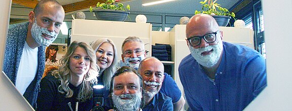 Gruppenfoto in einem Salon: Mehrere Erwachsene stehen dicht zusammen und lachen in einen Spiegel. Die meisten Männer tragen Spaß-Bärte aus Rasierschaum; zwei Frauen ohne Schaum posieren daneben. Fröhliche, lockere Stimmung.