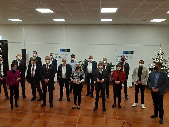 Gruppe von rund zwanzig Erwachsenen steht mit Abstand in einem Saal, alle tragen medizinische Masken. Im Hintergrund zwei Schilder mit der Aufschrift „Medizincampus Oberfranken“. Die Personen blicken zur Kamera, wirken wie bei einer offiziellen Veranstaltung oder Vorstellung.