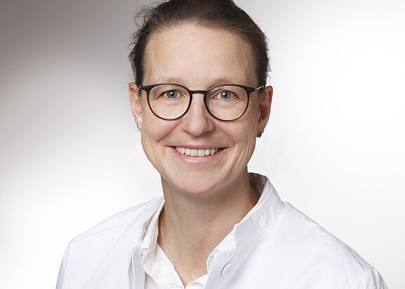 Porträt einer lächelnden Person mit Brille in weißem Arztkittel vor neutralem Hintergrund; Kopf-und-Schultern-Aufnahme, direkter Blick in die Kamera.