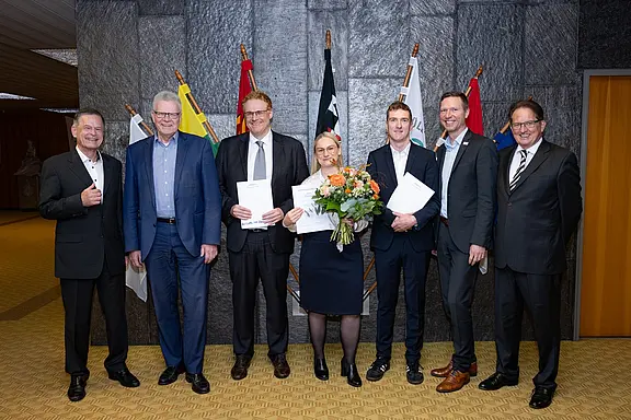 Eine Gruppe von sieben Personen steht in einem Raum, von denen einige Urkunden und eine Blumenstrauß in der Hand halten. Im Hintergrund sind Flaggen zu sehen, was auf eine offizielle Veranstaltung hinweist. Alle wirken erfreut und posieren für ein Foto.