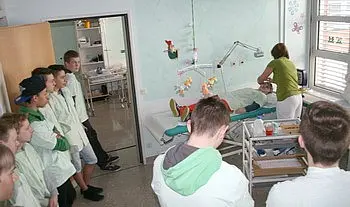 Eine Gruppe von Jugendlichen beobachtet einen medizinischen Eingriff oder eine Behandlung in einem Krankenhauszimmer. Eine Fachkraft kümmert sich um einen Patienten, der auf einem Krankenbett liegt. Die Umgebung ist sauber und praktisch gestaltet.