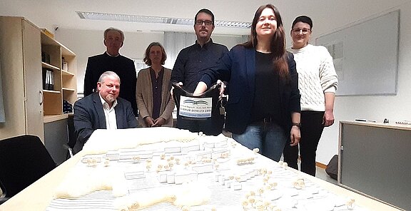 Sechs Personen stehen in einem Raum um ein großes, detailliertes Modell eines Geländes herum. Eine Person hält einen Behälter mit einer Aufschrift, die auf ein Projekt hinweist. Die anderen blicken in die Kamera, während sie das Modell betrachten.