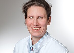 Dr. med. Sarah Prüfer Porträt einer lächelnden Frau in weißem Arztkittel vor hellem, neutralem Hintergrund. Sie hat dunkles, zurückgebundenes Haar und trägt kleine Ohrstecker. Professioneller, freundlicher Ausdruck.