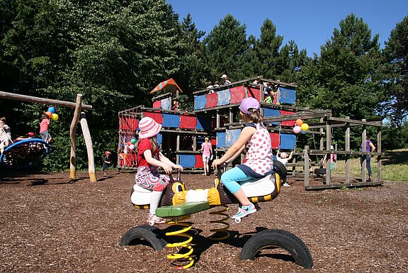 Auf einem sonnigen Spielplatz sitzen zwei Kinder auf einer Federwippe im Vordergrund; im Hintergrund klettern weitere Kinder auf bunten Holz- und Seilkonstruktionen.