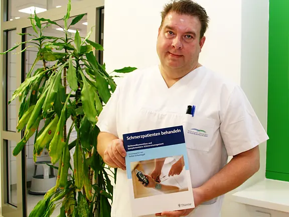 Pflegekraft in weißer Dienstkleidung hält ein Buch mit dem Titel „Schmerzpatienten behandeln“ und zeigt es in die Kamera. Das Cover zeigt eine medizinische Behandlung am Handrücken. Die Umgebung wirkt klinisch.