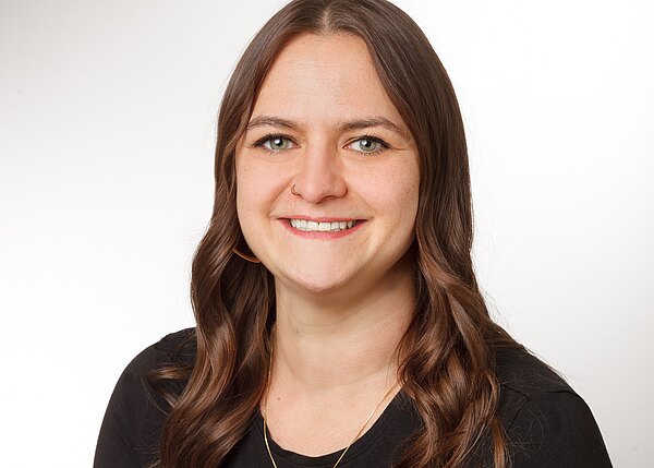 Porträt einer lächelnden Frau mit langen, gewellten braunen Haaren, die in die Kamera blickt. Neutraler Studiohintergrund, Oberkörper sichtbar, schwarzes Oberteil und dezenter Schmuck.