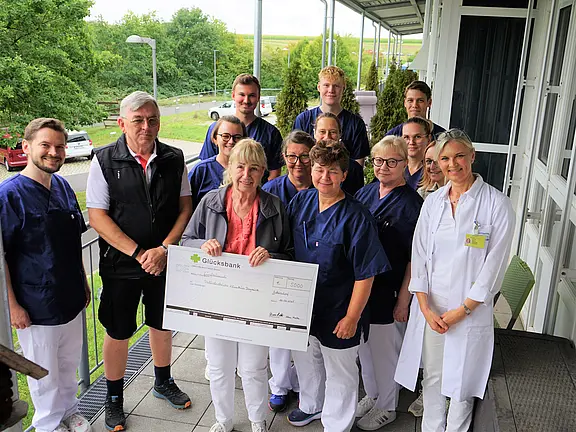 Die Bildbeschreibung zeigt eine Gruppe von Personen, darunter medizinisches Personal, die zusammen für ein Foto posieren. Eine Frau in der Mitte hält einen großen Scheck mit der Aufschrift „Glücksbank“. Alle haben freundliche Ausdrücke und stehen auf einem Balkon mit grünem Hintergrund.