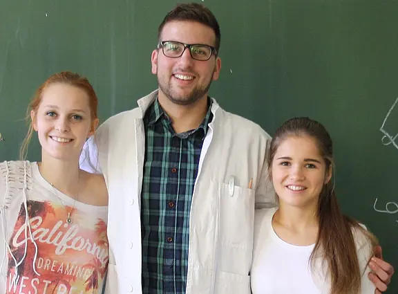 Vor einer Tafel stehen drei junge Menschen nah beieinander und lächeln. Die mittlere Person trägt Brille und weißen Laborkittel, die beiden anderen Freizeitkleidung. Sie legen einander die Arme um die Schultern, wirken freundlich und verbunden.