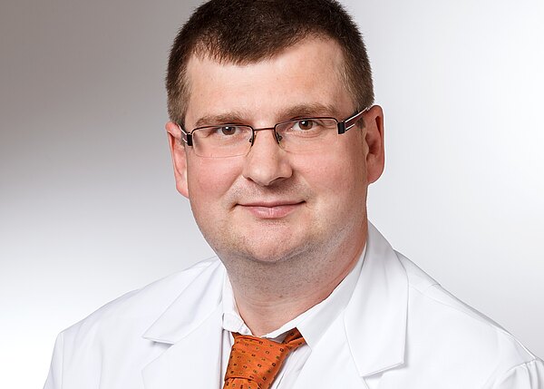 Porträt eines Mannes mit Brille in weißem Arztkittel und orangefarbener Krawatte. Er lächelt leicht, Blick zur Kamera, neutraler Hintergrund.