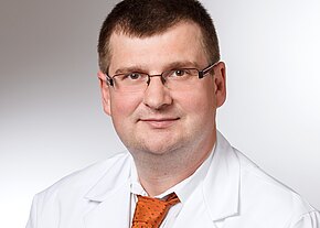 Porträt eines Mannes mit Brille in weißem Arztkittel und orangefarbener Krawatte. Er lächelt leicht, Blick zur Kamera, neutraler Hintergrund.