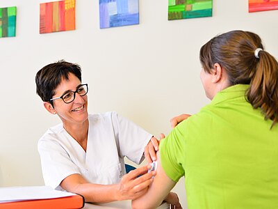 Eine Ärztin in einem weißen Kittel lächelt und verabreicht einer Patientin, die ein grünes Shirt trägt, eine Injektion in den Arm. Im Hintergrund sind farbige Kunstwerke an der Wand zu sehen.