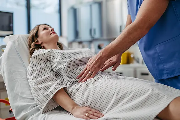 Eine Frau liegt in einem Krankenhausbett und trägt ein Krankenhaushemd. Ein männlicher Arzt untersucht sanft ihren Bauch, während sie ruhig aussieht. Im Hintergrund sind medizinische Geräte sichtbar, was auf eine medizinische Umgebung hinweist.
