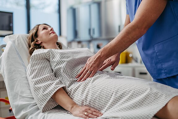 Eine Frau liegt in einem Krankenhausbett und trägt ein Krankenhaushemd. Ein männlicher Arzt untersucht sanft ihren Bauch, während sie ruhig aussieht. Im Hintergrund sind medizinische Geräte sichtbar, was auf eine medizinische Umgebung hinweist.