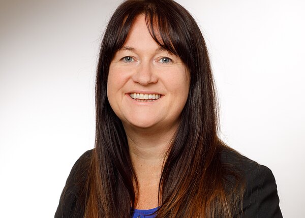 Porträt einer lächelnden Frau mit langen dunkelbraunen Haaren. Sie blickt in die Kamera, trägt ein dunkles Jackett und ein blaues Oberteil. Heller, neutraler Hintergrund; professionelles Business-Foto.