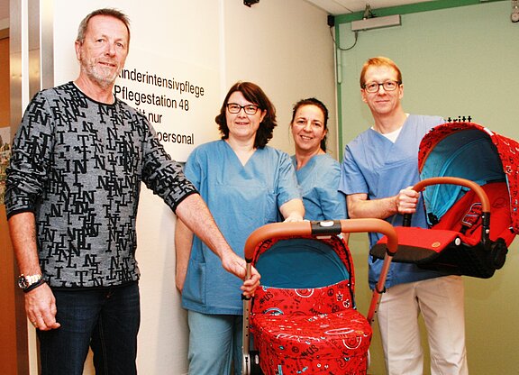 Ein Mann und drei Pflegekräfte in blauen Arbeitskleidung stehen neben einem Kinderwagen und einer Tragetasche für Babys. Im Hintergrund ist ein Schild mit der Aufschrift „Kindernintensivpflege, Pflegeestation 48, nur für Personal“ sichtbar.