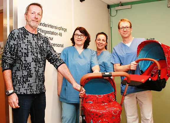 Ein Mann und drei Pflegekräfte in blauen Arbeitskleidung stehen neben einem Kinderwagen und einer Tragetasche für Babys. Im Hintergrund ist ein Schild mit der Aufschrift „Kindernintensivpflege, Pflegeestation 48, nur für Personal“ sichtbar.
