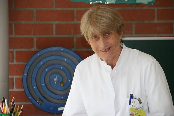 Ältere Frau in weißem Kittel lächelt freundlich in die Kamera. Sie wirkt wie medizinisches Personal. Fokus auf Gesicht und Oberkörper; neutrale Innenraumkulisse.