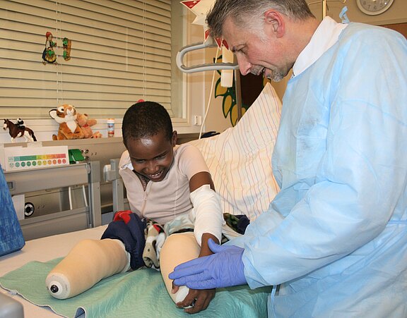 Im Krankenhausbett sitzt ein lächelndes Kind mit bandagierten Beinstümpfen und verbundenem Arm. Ein Arzt in Schutzkittel und Handschuhen unterstützt behutsam die Beine. Die Szene zeigt fürsorgliche medizinische Versorgung und Hoffnung.