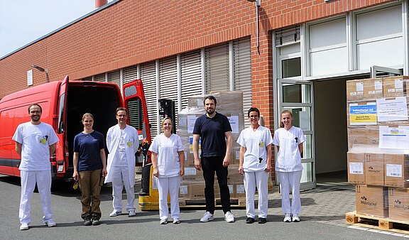 Mehrere Pflegekräfte und Helfer stehen vor einem Gebäude neben einem roten Lieferwagen und Paletten mit Kartons. Szene zeigt die Anlieferung von medizinischen Materialien oder Ausrüstung.
