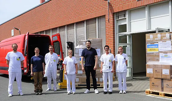 Mehrere Pflegekräfte und Helfer stehen vor einem Gebäude neben einem roten Lieferwagen und Paletten mit Kartons. Szene zeigt die Anlieferung von medizinischen Materialien oder Ausrüstung.