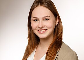 Freundliches Porträt: junge Frau mit langen, rötlich-braunen Haaren, leicht schräg zur Kamera, lächelt. Sie trägt ein beiges Sakko über weißem Top, kleine Ohrstecker. Neutraler, heller Hintergrund, professioneller Eindruck.