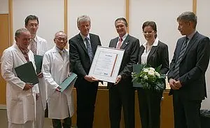 Gruppenfoto von fünf Personen in Anzug, die eine Urkunde halten, umgeben von medizinischem Personal in Kitteln. Sie stehen in einem Raum mit hellen Wänden, einige Personen halten Dokumente und eine Blumenstrauß wird überreicht. Die Stimmung ist feierlich und anerkennend.