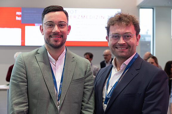 Zwei Männer in Anzügen mit blauen Konferenzlanyards stehen nebeneinander und lächeln in die Kamera. Hinter ihnen ein heller Messestand mit unscharfem Bildschirm und mehreren Personen im Hintergrund. Fokus auf freundliches Porträt während einer Fachveranstaltung.