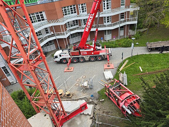 Ansicht von oben einer Baustelle: Ein großer roter Autokran mit ausgefahrenen Stützen arbeitet an einem Turmdrehkran; mehrere Kranteile liegen abgesperrt am Boden im Hof eines Backsteingebäudes.