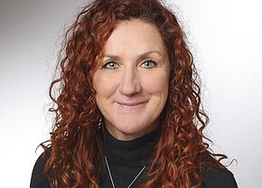 Sylvia Scharf Porträt einer Frau mit lockigem, rotem Haar und einem freundlichen Lächeln. Sie trägt einen schwarzen Rollkragenpullover und hat grüne Augen. Der Hintergrund ist hell und neutral, wodurch ihr Gesicht im Fokus steht.