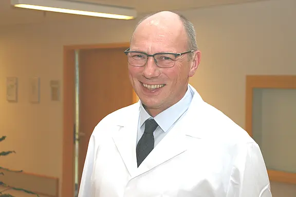 Ein lächelnder Mann mit Brille trägt weißen Kittel und Krawatte, steht in einem hellen Klinikflur und blickt in die Kamera.